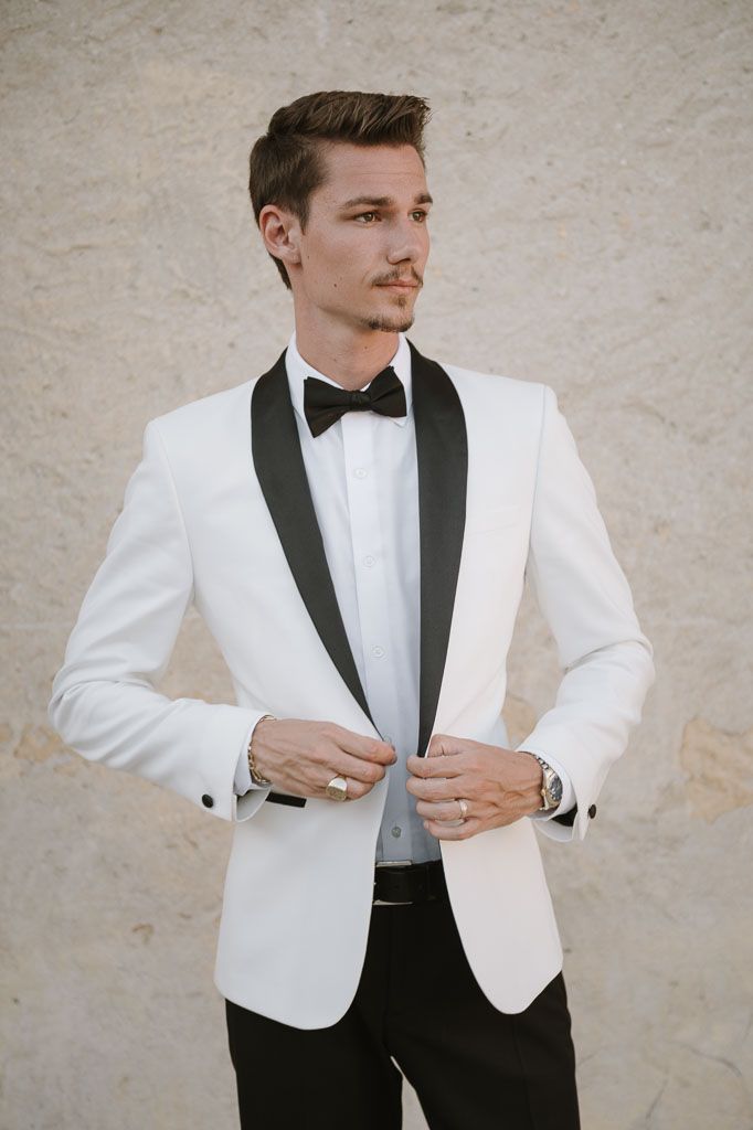 A groom wearing a white tuxedo jacket with black lapels and bow tie adjusts his cuff.