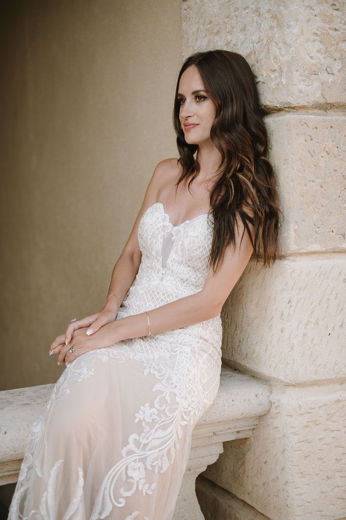 A bride in a fitted lace wedding gown with sheer details poses elegantly against a stone wall.