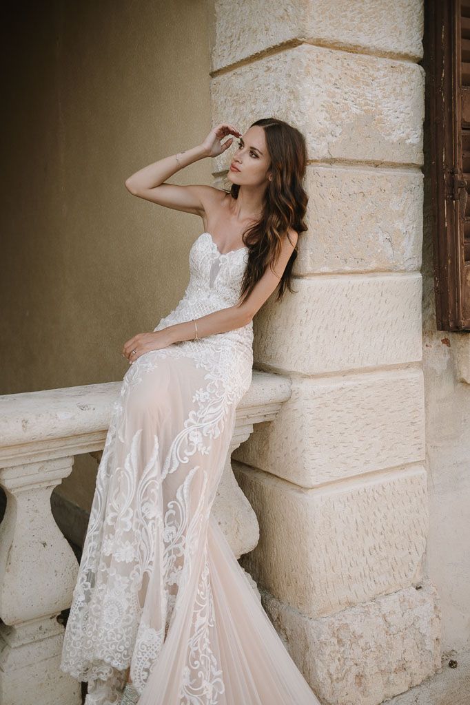 A bride in a sheer lace wedding dress poses gracefully on stone balustrade.