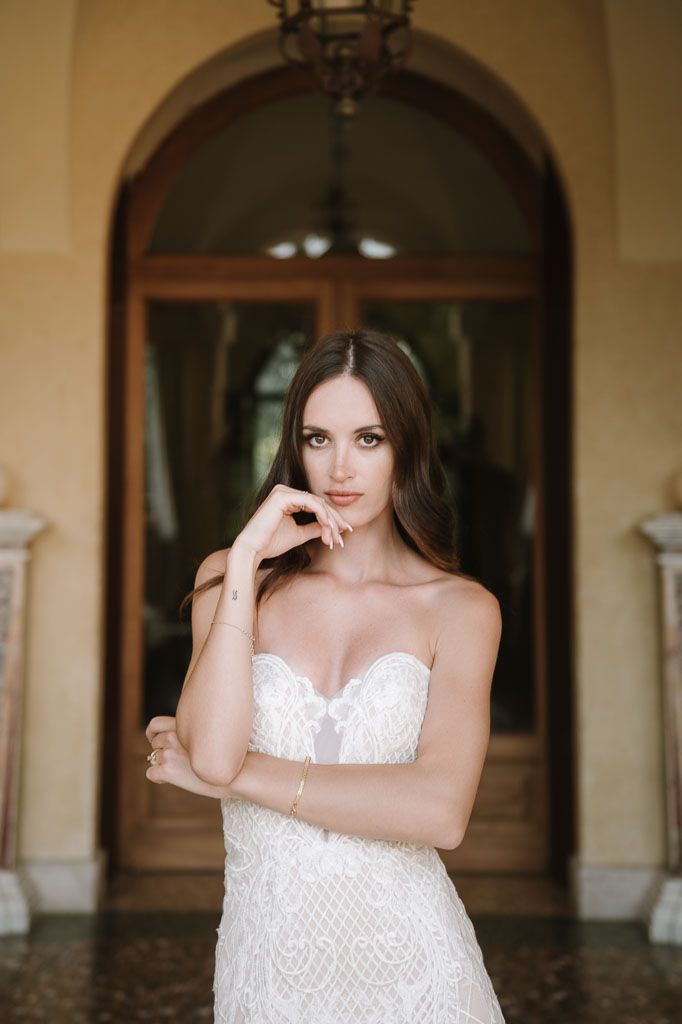 Bride in strapless lace gown poses in front of arched doorway with classic architecture.