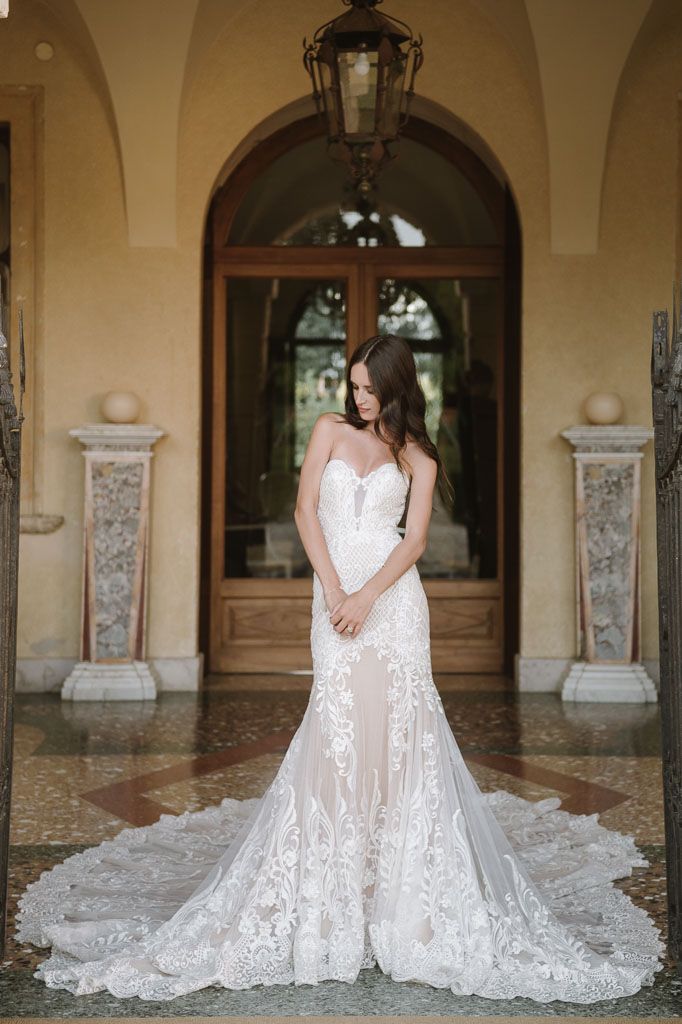 Bride in fitted lace gown with cathedral train stands before arched doorway.