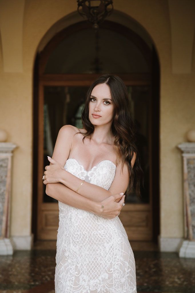 A strapless white lace wedding dress is showcased in an elegant archway setting with warm lighting.