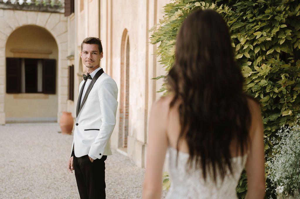 A stylish wedding couple poses in front of an elegant villa with green foliage.