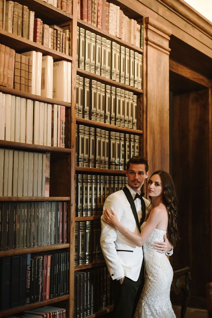 Elegant couple in formal attire embraces in a vintage library setting with floor-to-ceiling bookshelves.