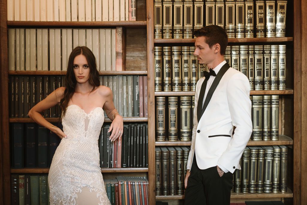 Stylish couple in white and black formal wear pose separately in an antique library setting.