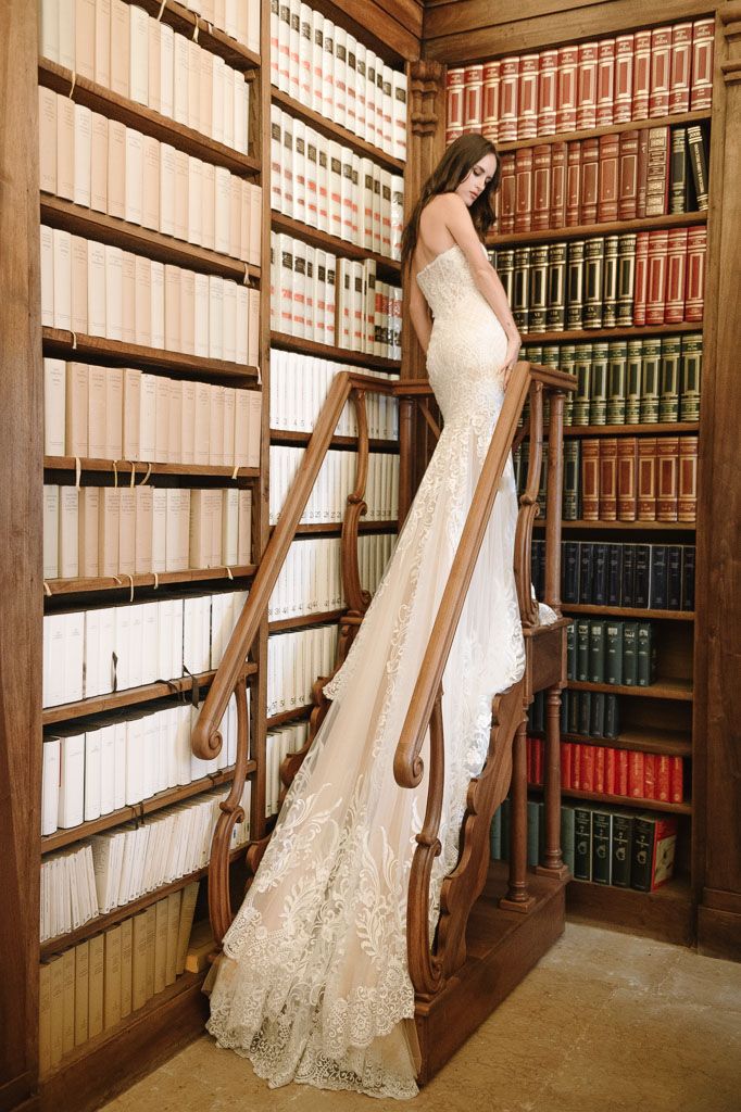 Woman in fitted lace wedding dress poses on wooden library ladder among historic book collection.