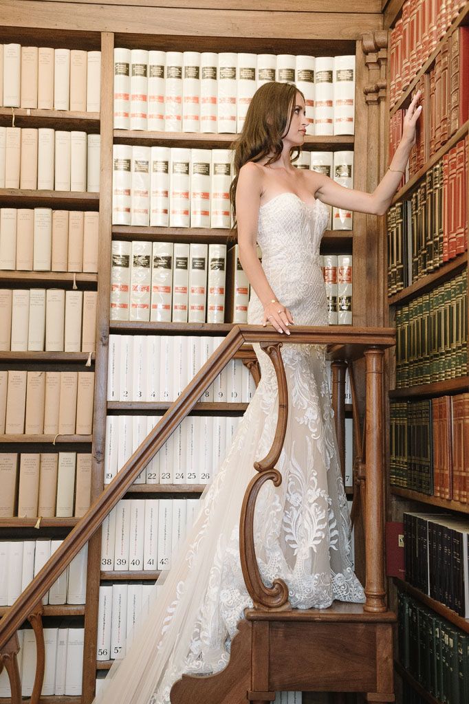 Bride in strapless wedding gown poses gracefully on ornate library ladder among bookshelves.