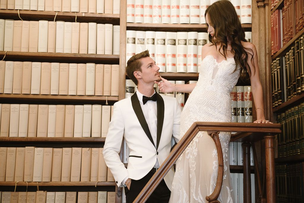 Couple shares playful moment on library ladder with groom in white tuxedo looking up at bride.