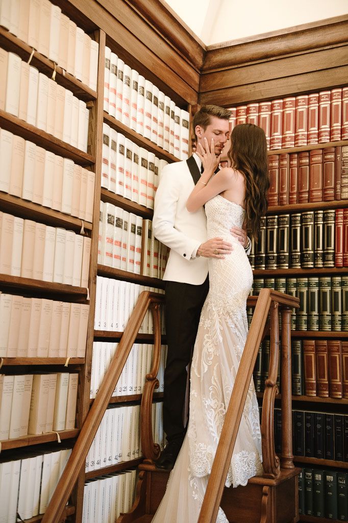 Romantic couple embraces on library ladder surrounded by floor-to-ceiling bookshelves.