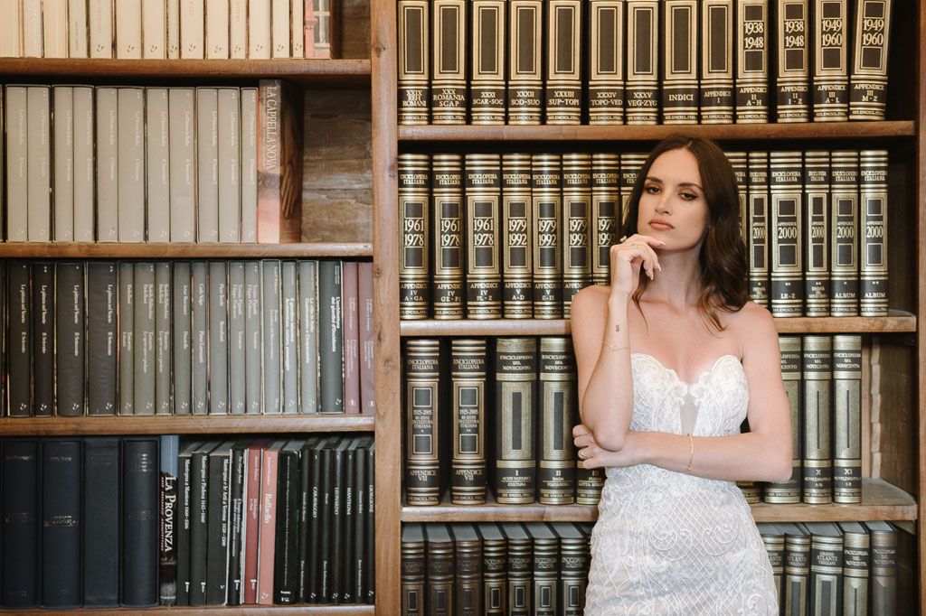 Bride in lace gown poses thoughtfully against vintage book collection in classic library.