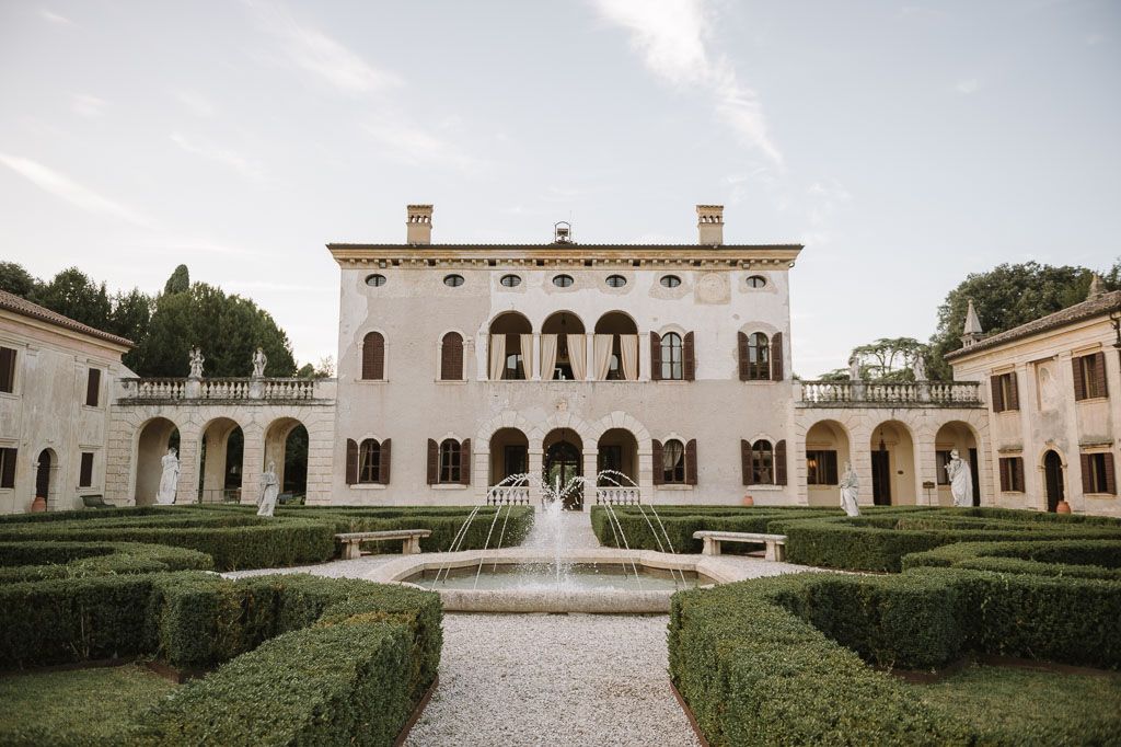 Elegant Italian villa with white facade, arched windows and manicured hedge garden with central fountain.