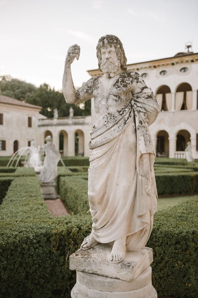 Classical stone statue in draped robes standing among formal garden hedges.