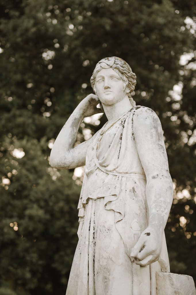 Classical marble statue of draped figure against blurred garden background.
