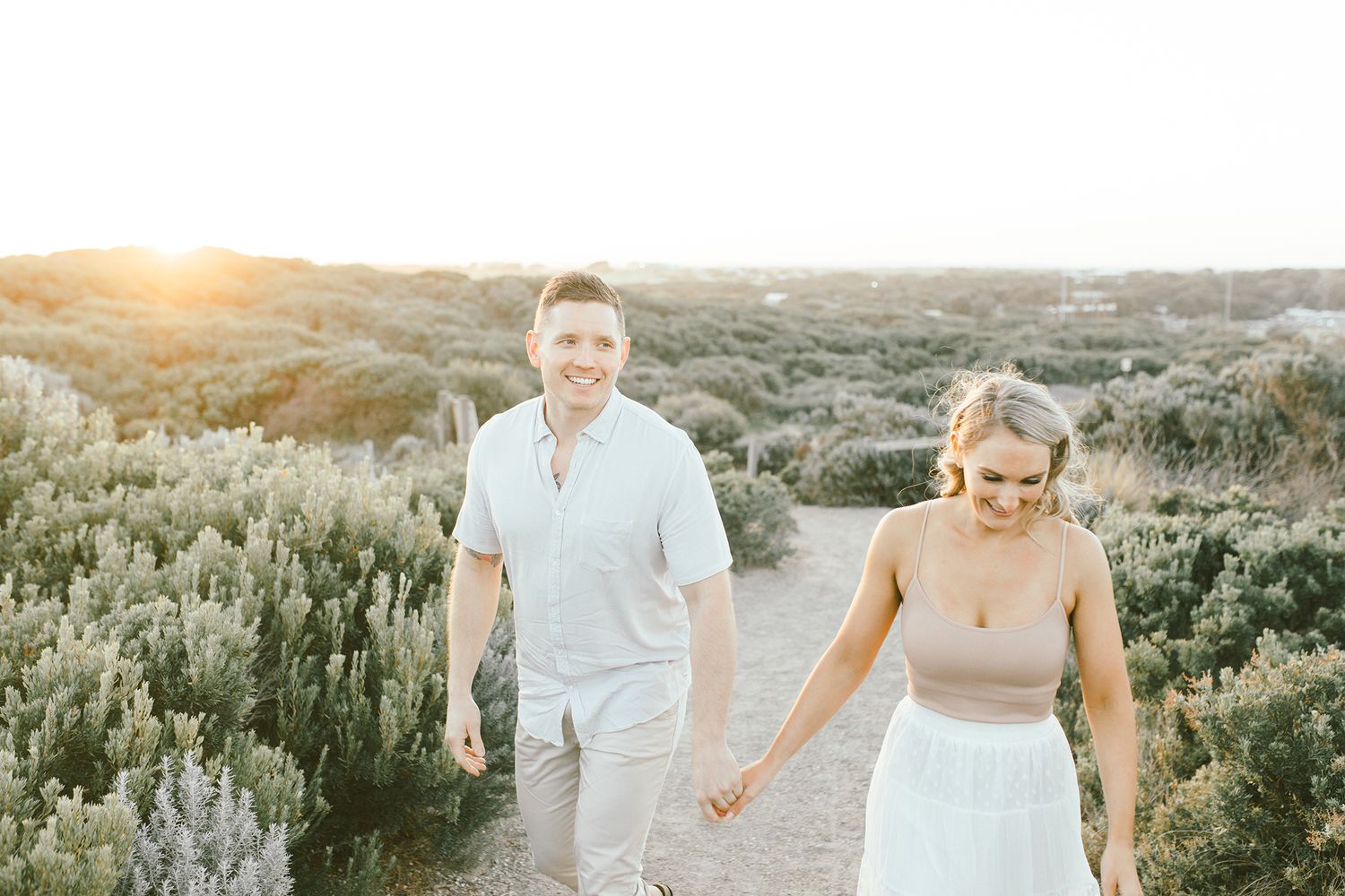 A couple walks hand in hand on a nature trail during golden hour among sage brush and desert vegetation.