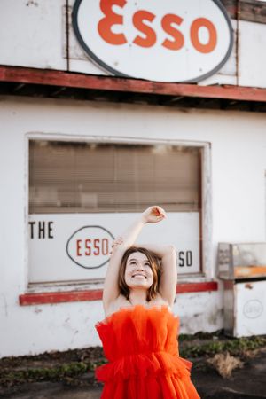 Vibrant red tulle dress contrasts against vintage Esso gas station signage in urban setting.