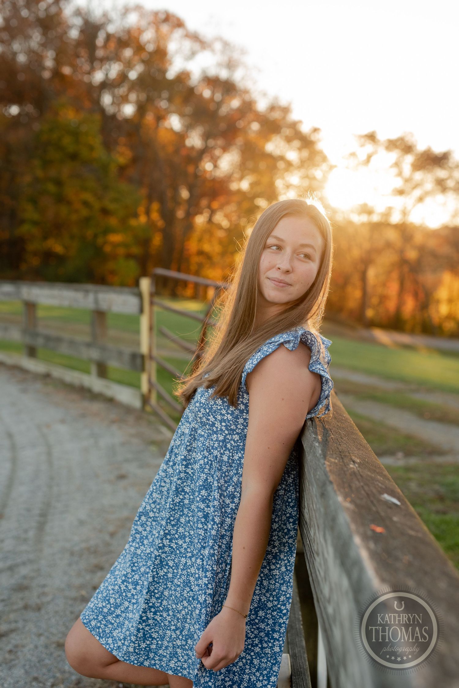 A blue floral dress contrasts against autumn scenery along a curved fence path.