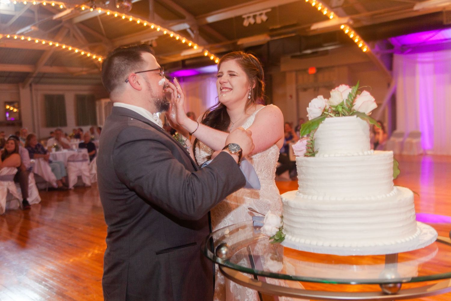 Elegant wedding reception venue with string lights and purple accents during cake cutting moment.