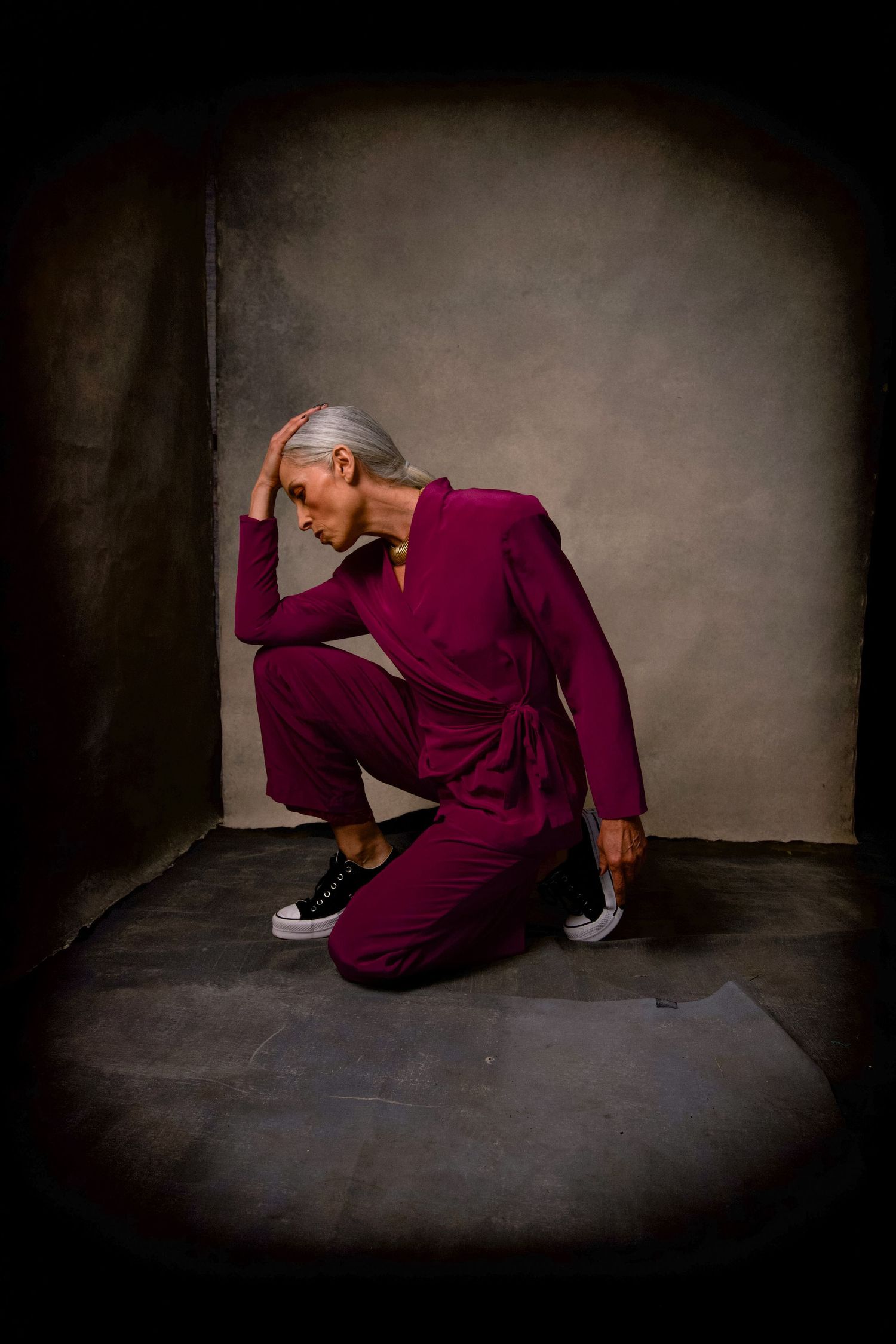 Editorial image of figure in a burgundy jumpsuit crouches dramatically in a moody dark corner setting.