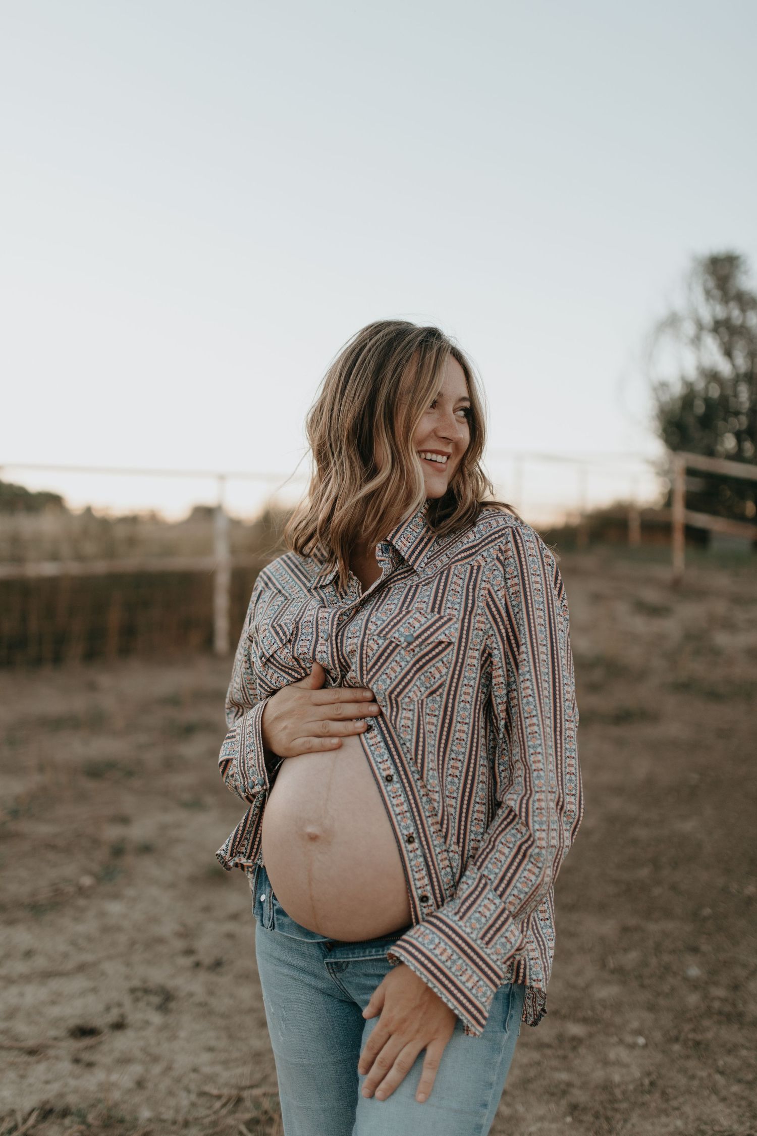 An expectant mother in a patterned top touches her belly while smiling in warm evening light.