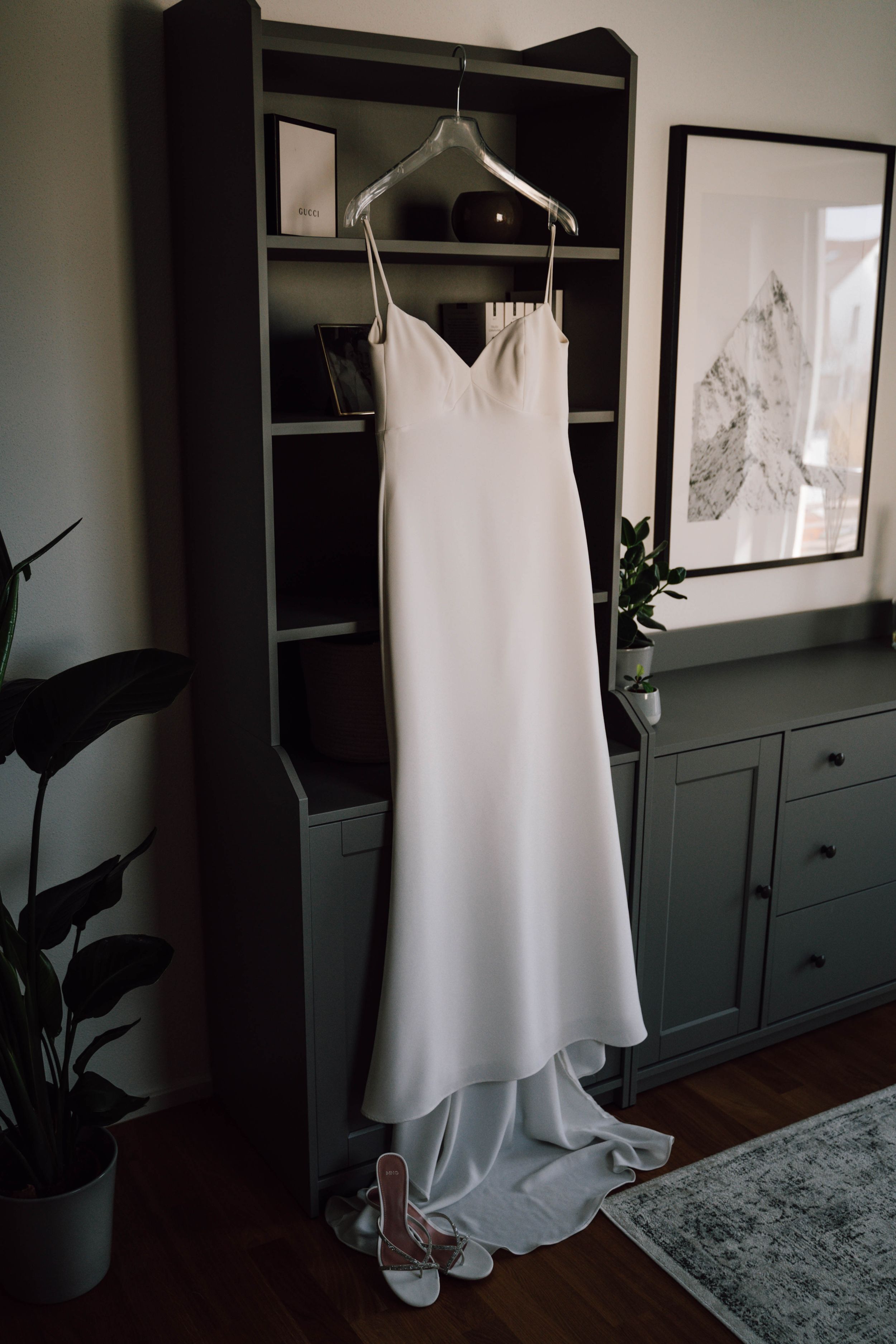 White wedding gown displayed on a hanger in a room with dark walls and built-in storage.