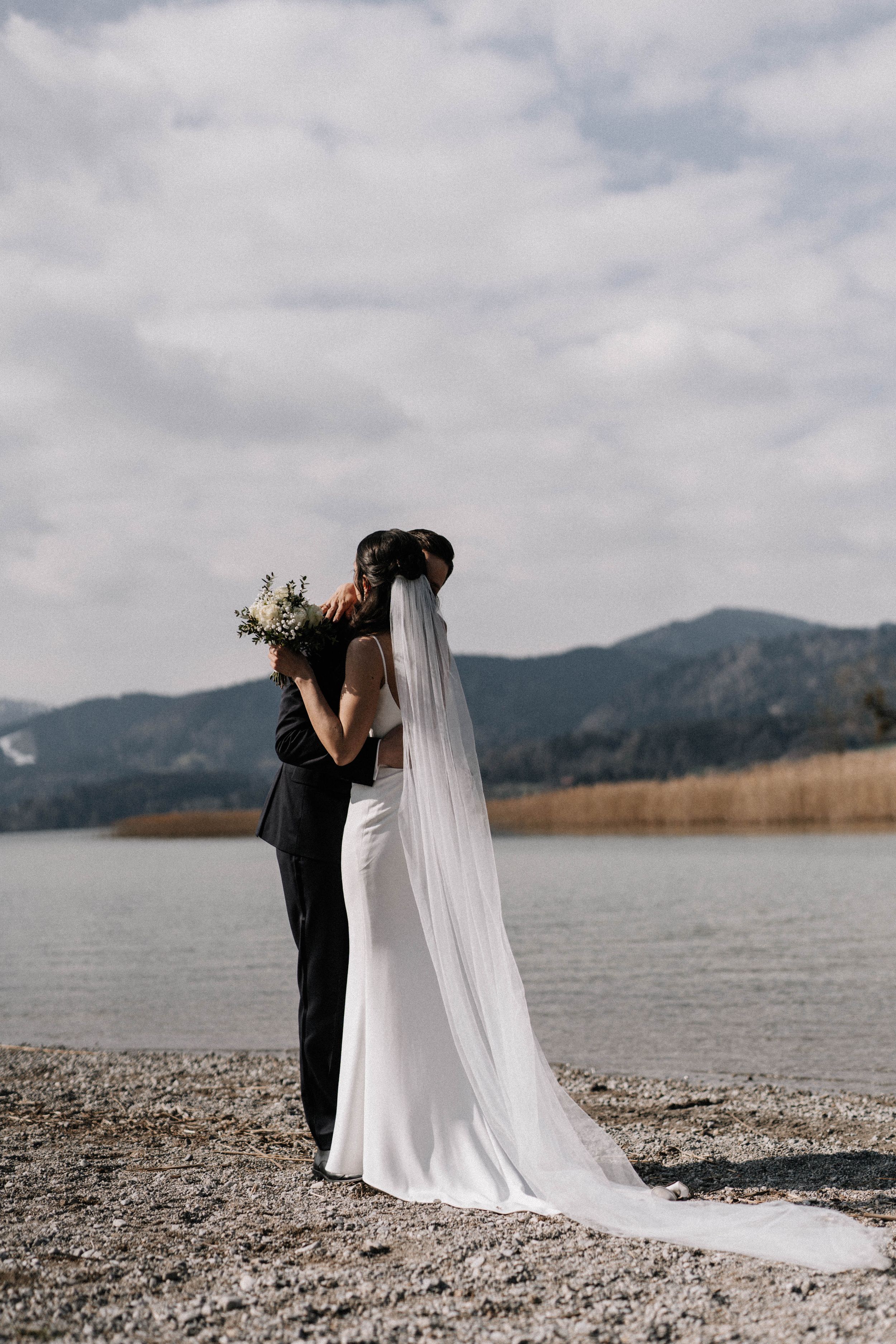 Couple embracing on a rocky shoreline with mountains and lake in the background.