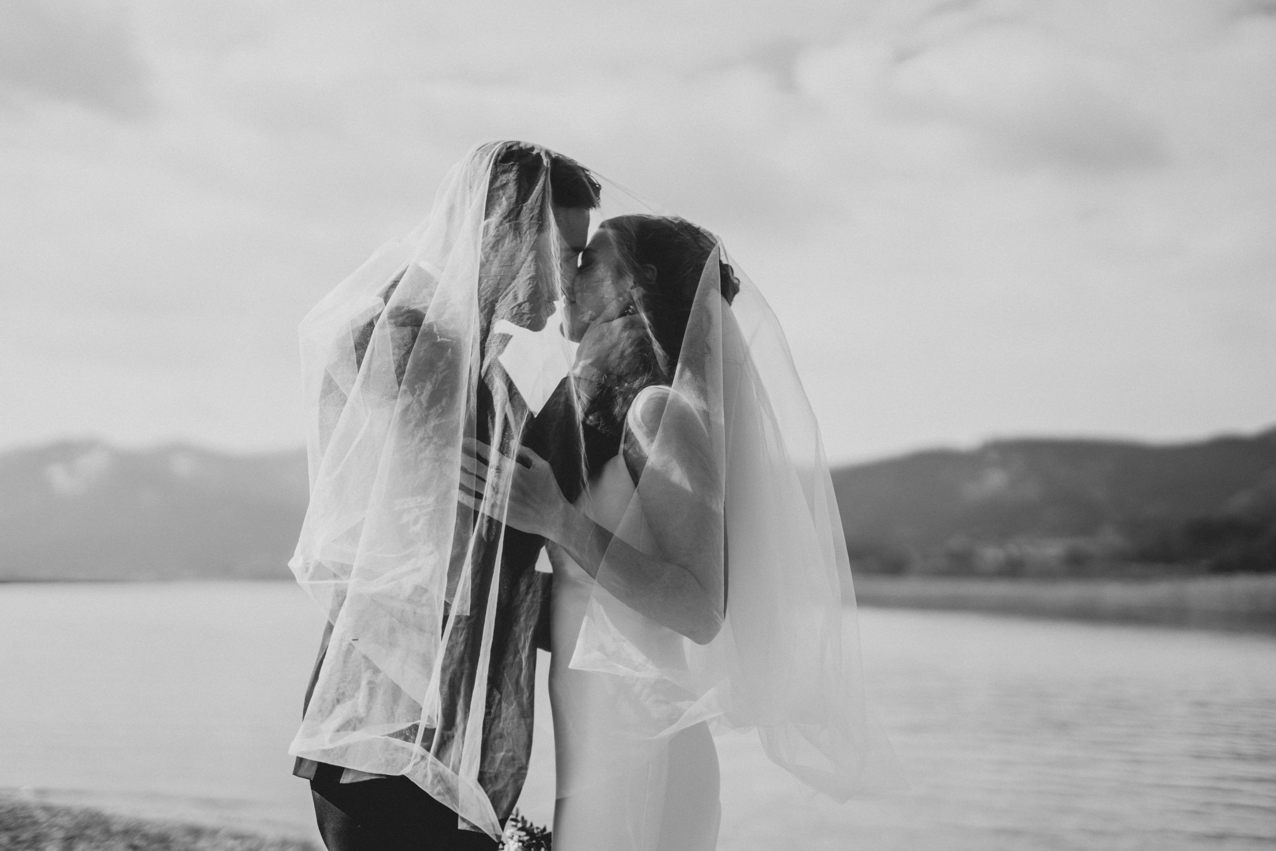 A romantic wedding photoshoot with a couple sharing intimate moments under a flowing veil by a lake.