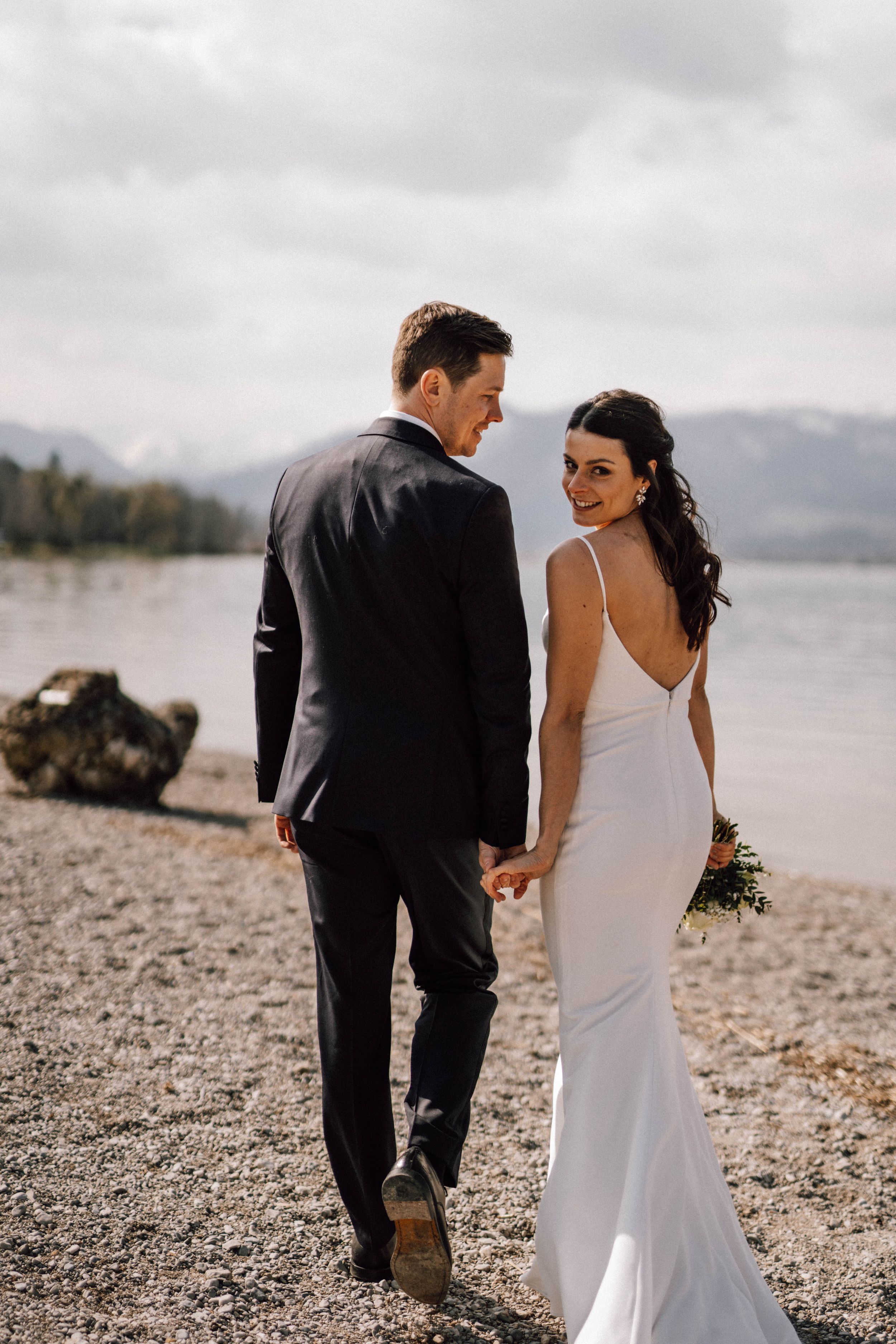 Newlyweds walking along a rocky beach with mountains in the background.