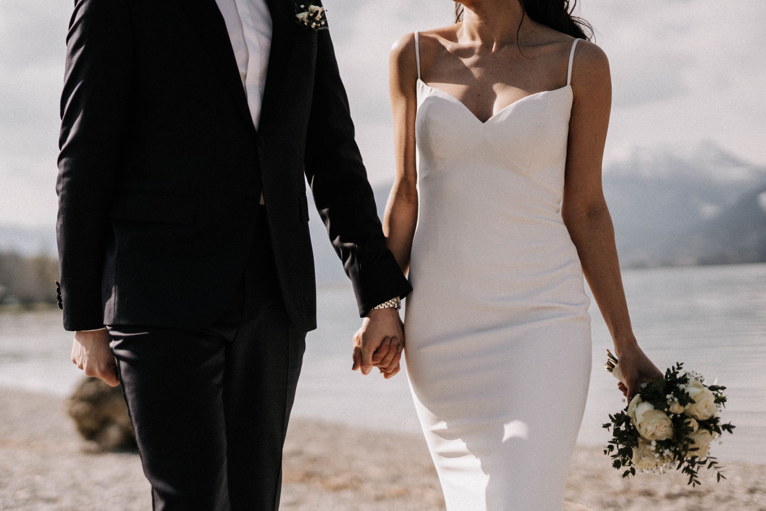 Wedding couple walking hand in hand on a pebble beach with mountain views.