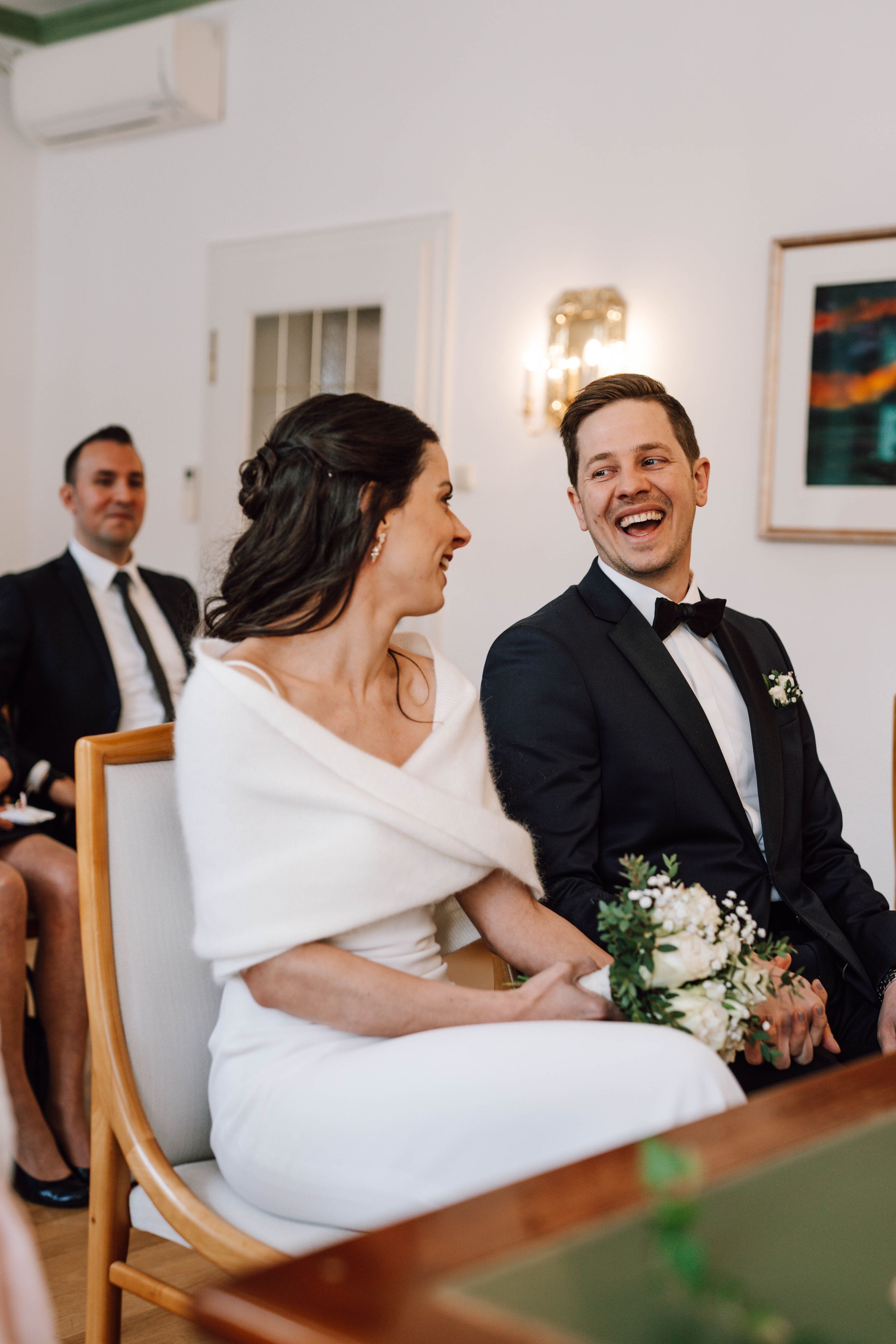 A joyful wedding ceremony moment captured indoors with the couple sharing laughter while seated.