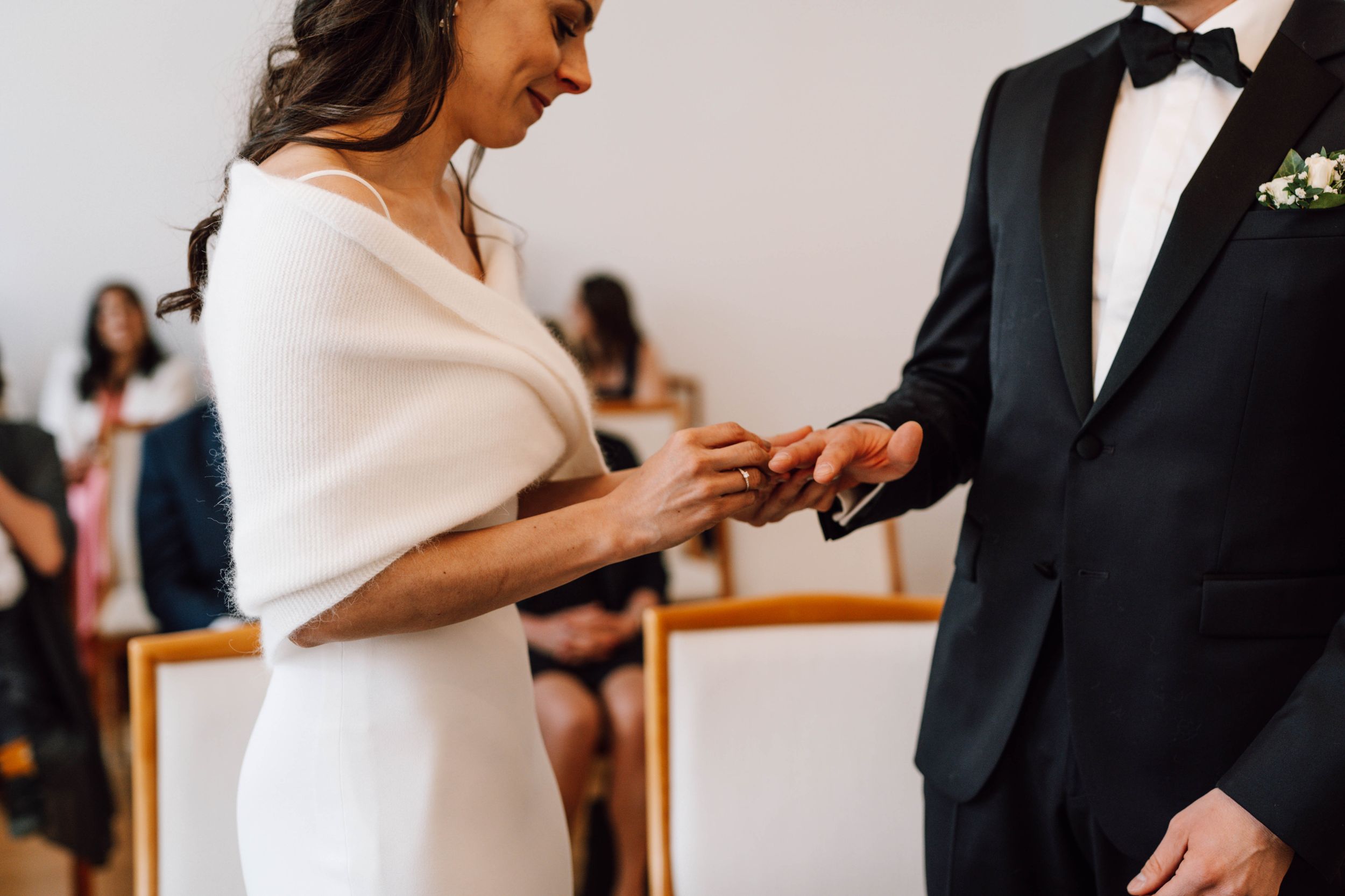 A wedding ceremony scene showing ring exchange between couple in elegant white dress and black tuxedo.