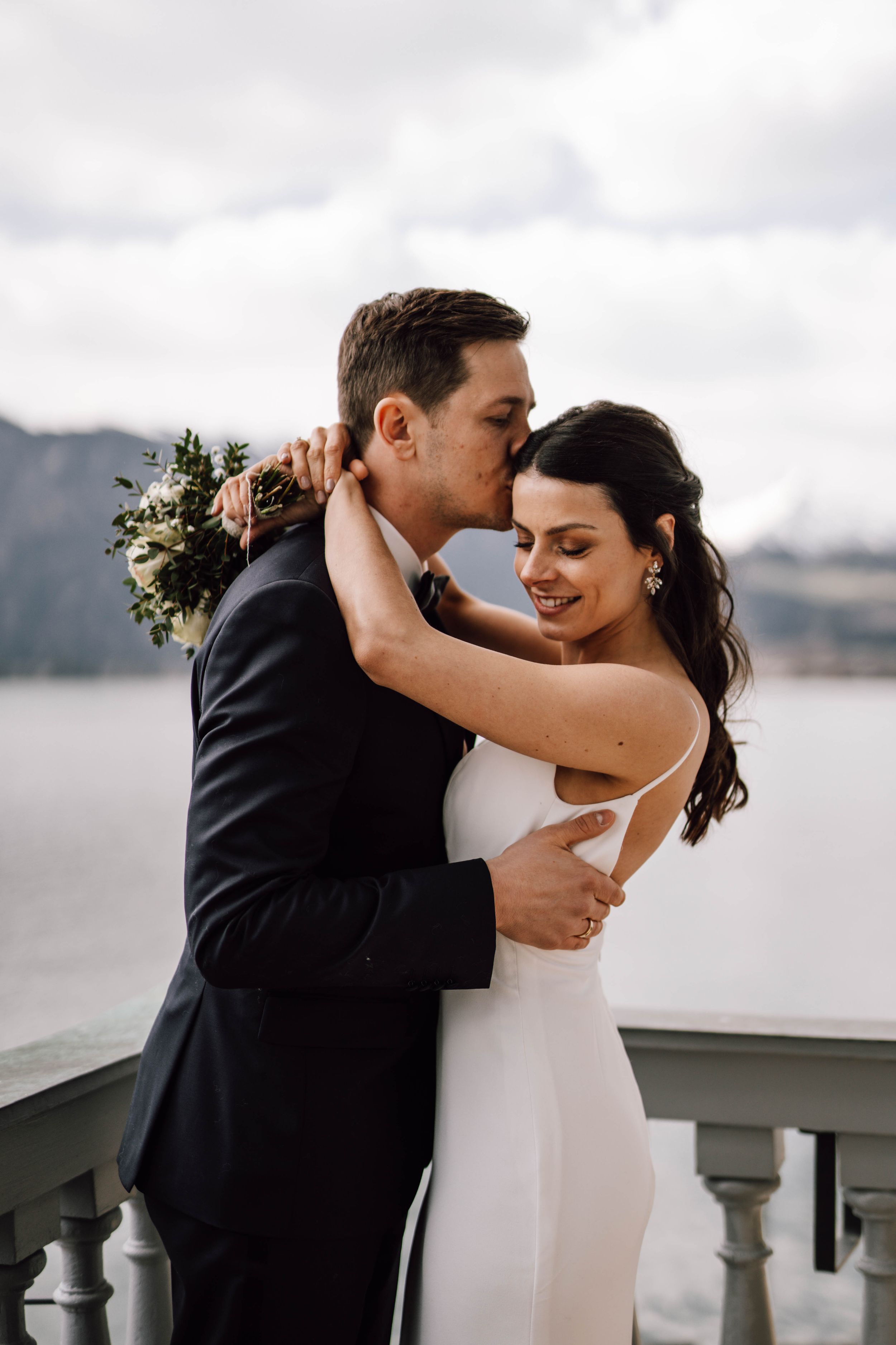 A romantic wedding portrait on a balcony overlooking mountains with couple in elegant attire embracing.