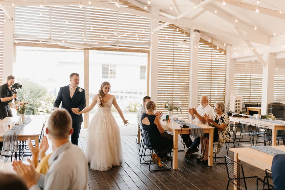 Newlyweds walk hand-in-hand into sunlit wedding reception venue with string lights and wooden slat walls.