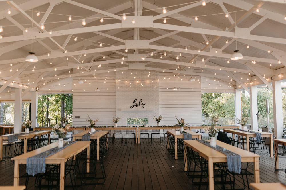 Elegant wedding reception venue with string lights, wooden beams, long farm tables, and black chairs on dark hardwood floors.