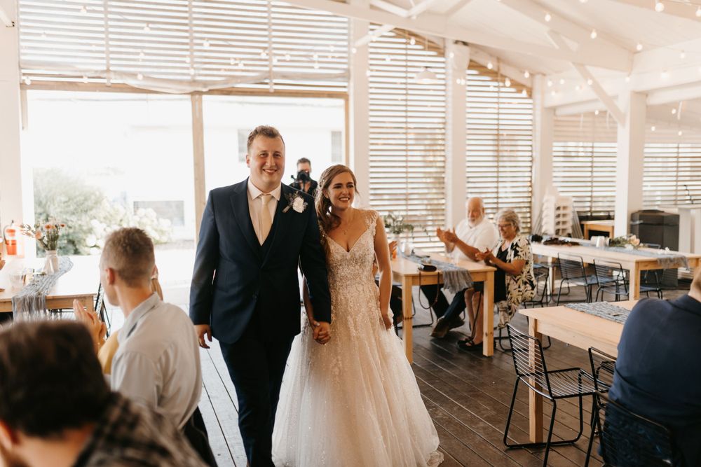 Smiling bride in lace gown and groom in navy suit enter bright, modern wedding reception venue together.