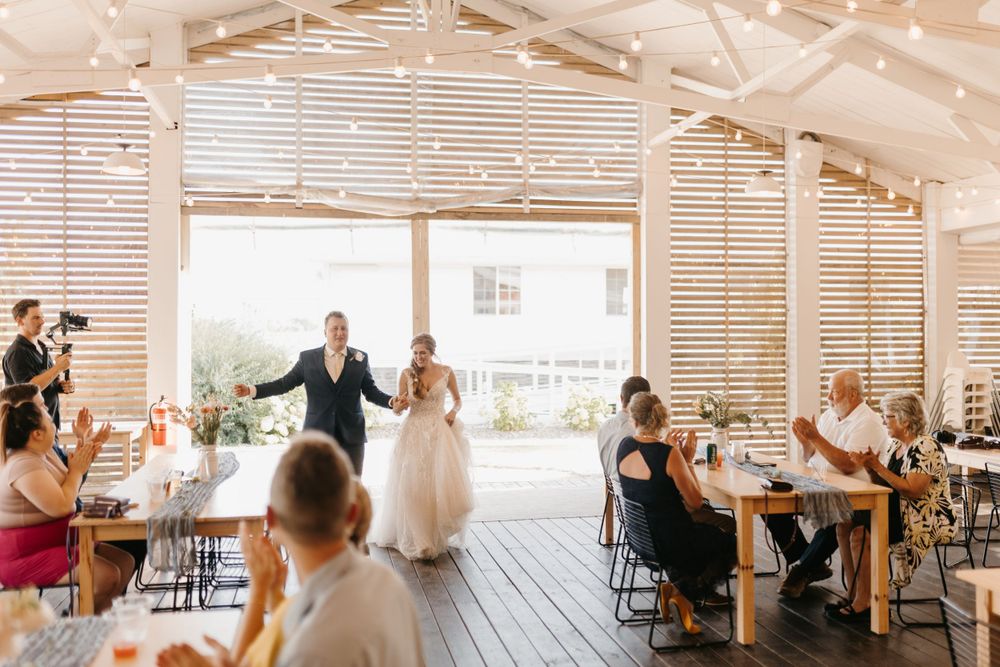 Bride and groom make grand entrance into rustic wedding reception hall as guests applaud and celebrate.