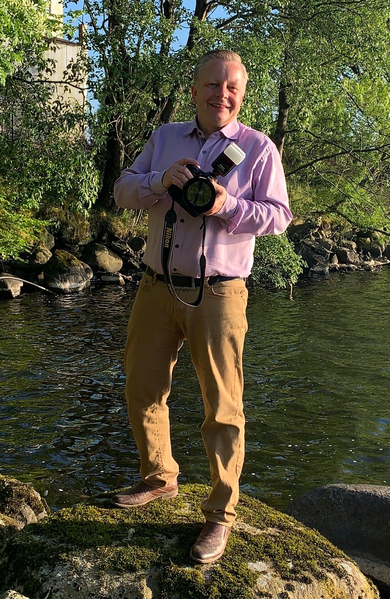 Smiling man holding a professional camera standing on rocks by a river outdoors.