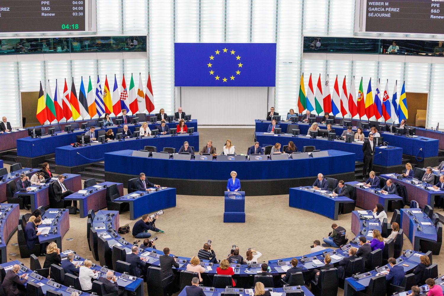 Interior view of European Parliament hemicycle chamber during session with EU flag and member state flags displayed.