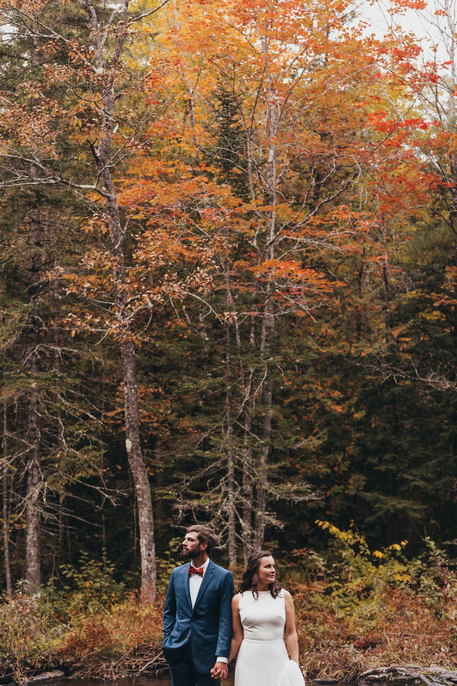 Newlyweds walk through vibrant autumn forest with orange and red foliage.
