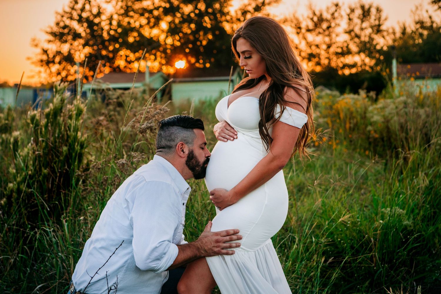 A couple shares an intimate moment in a field at sunset, with the person kissing a pregnant belly in a white dress.