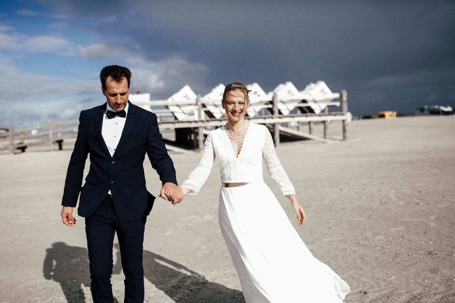 A couple on their wedding day walks hand in hand along a beachfront with modern buildings in the background.