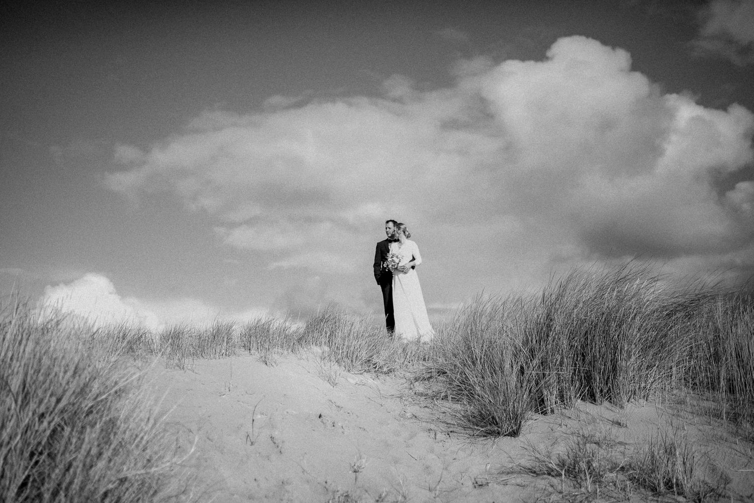 A romantic black and white wedding photograph capturing a couple standing atop a sand dune against dramatic clouds.