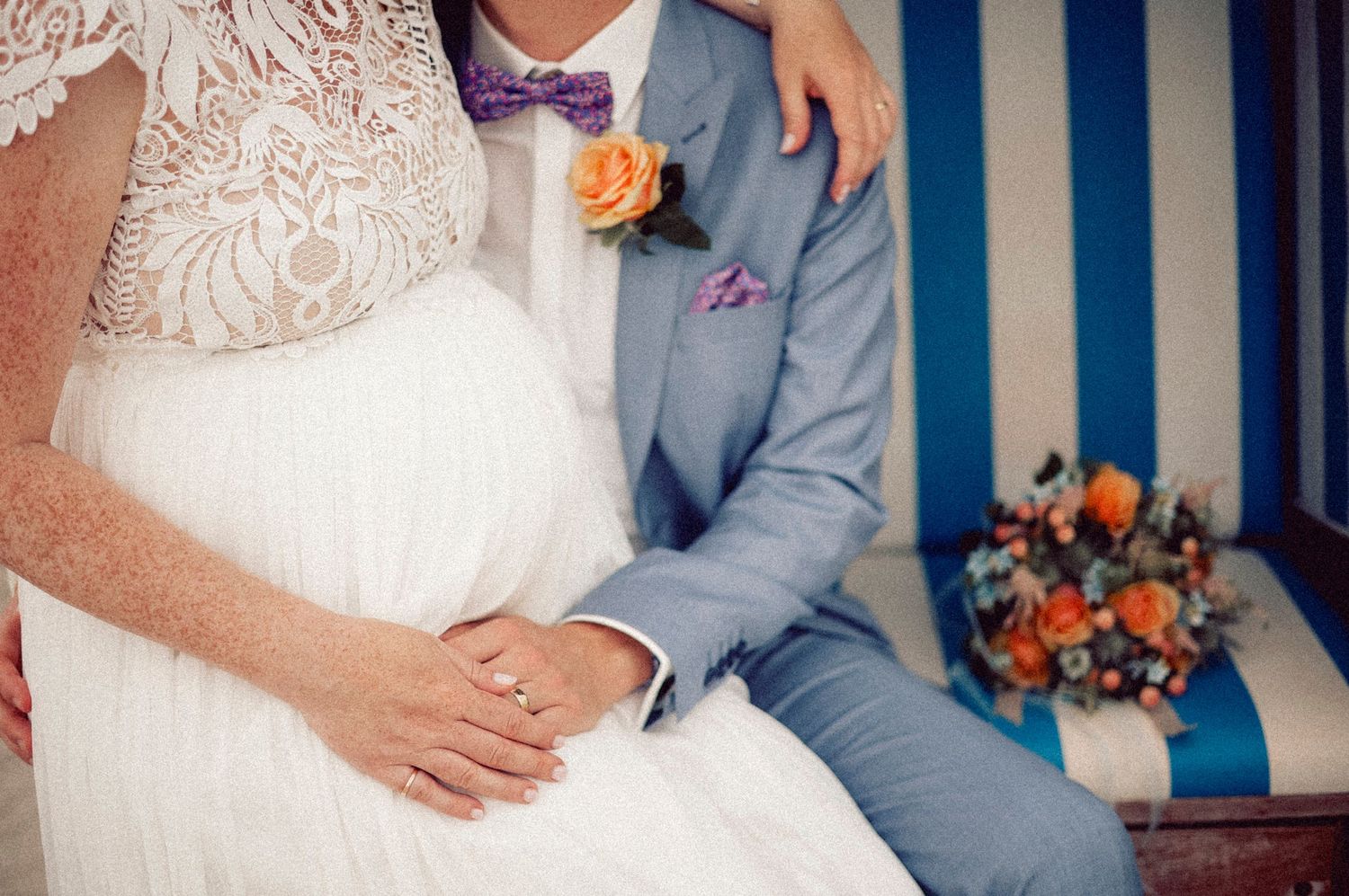 A bride in a white lace gown sits next to a groom wearing a light gray suit with coral boutonniere and blue bow tie.