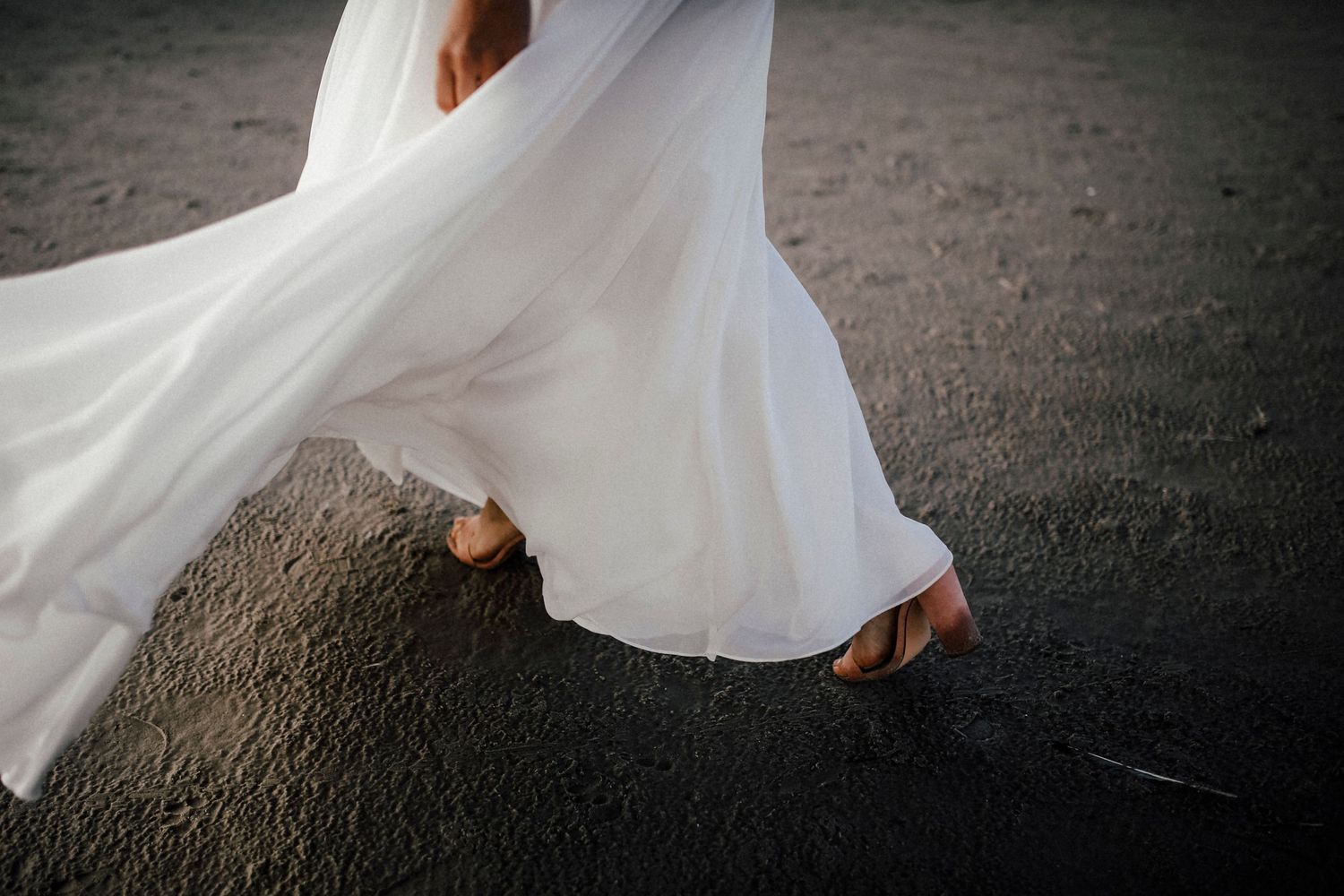 Close up of white wedding dress flowing in the wind with bride's feet peeking out on dark ground.