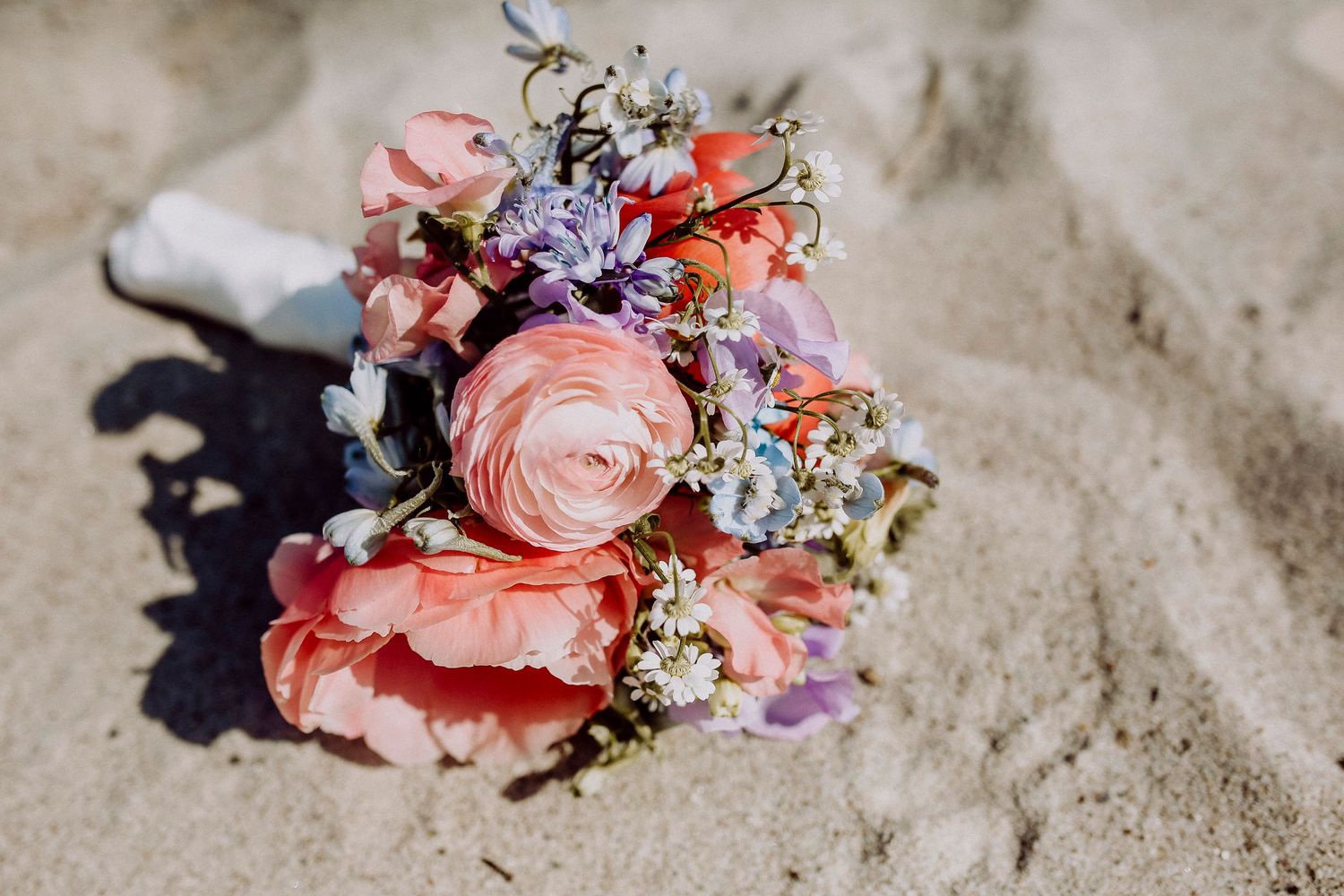 A delicate wedding bouquet with pink roses and small purple flowers rests on sandy beach in bright sunlight.