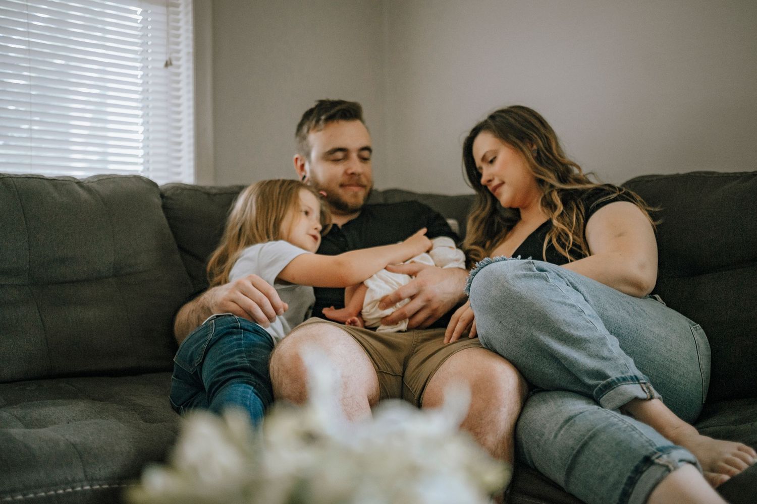 Family relaxes together on grey couch with newborn baby and young child in intimate home setting.