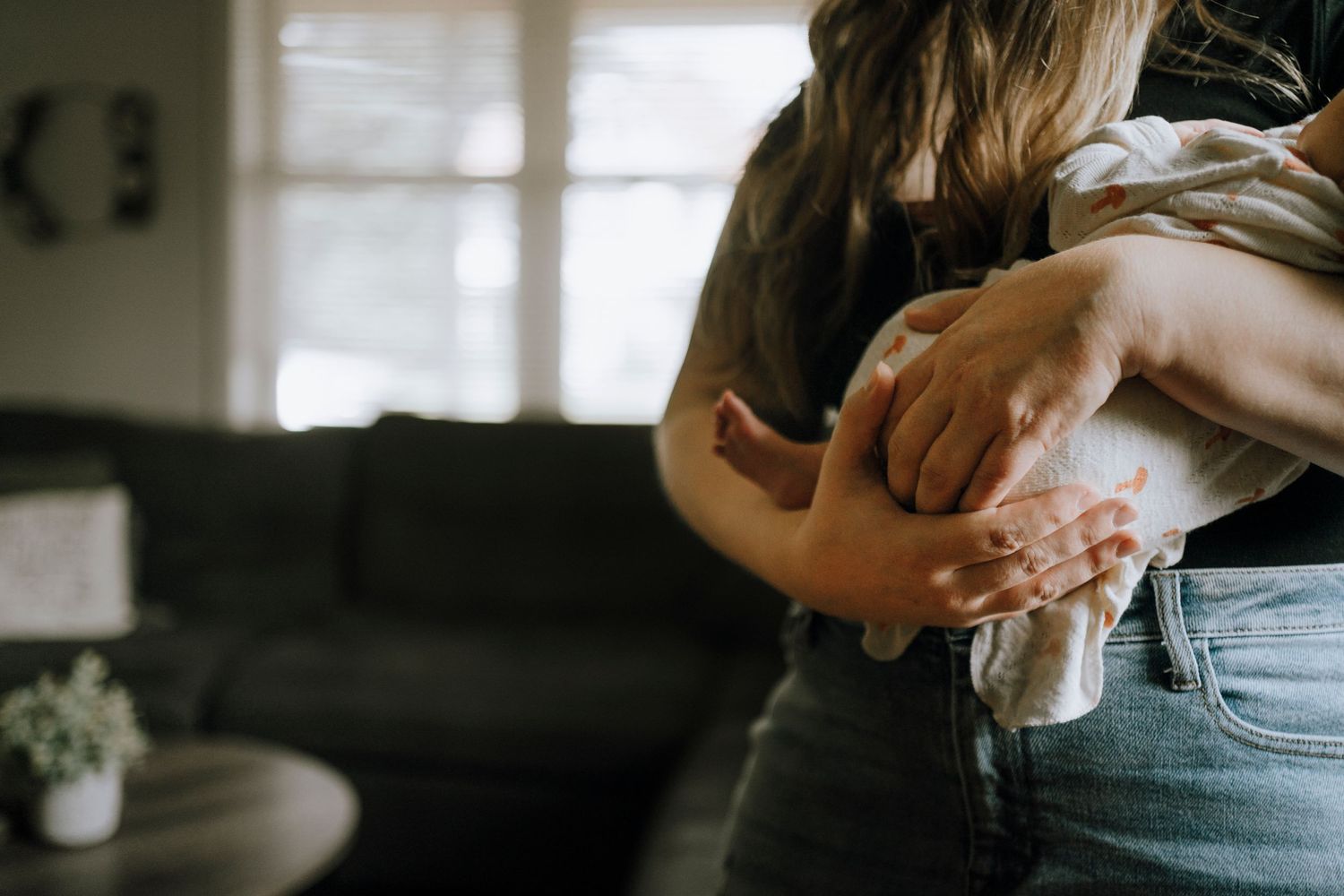 Parent holding newborn baby against soft window light in casual home setting.