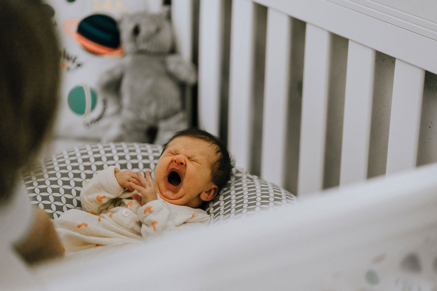 Baby yawning while lying in white crib with stuffed animals visible in background.