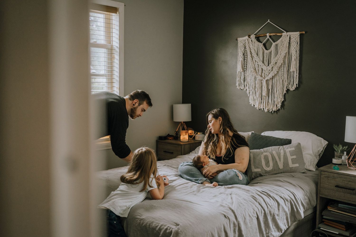 Family gathered on grey bed with macrame wall hanging in background.