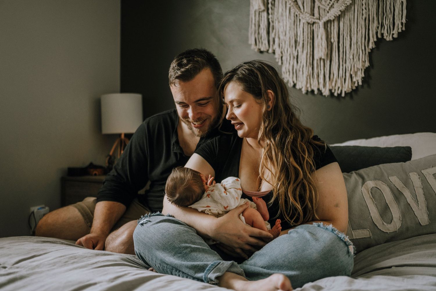 A couple relaxes on a bed with their newborn baby under a macrame wall hanging.