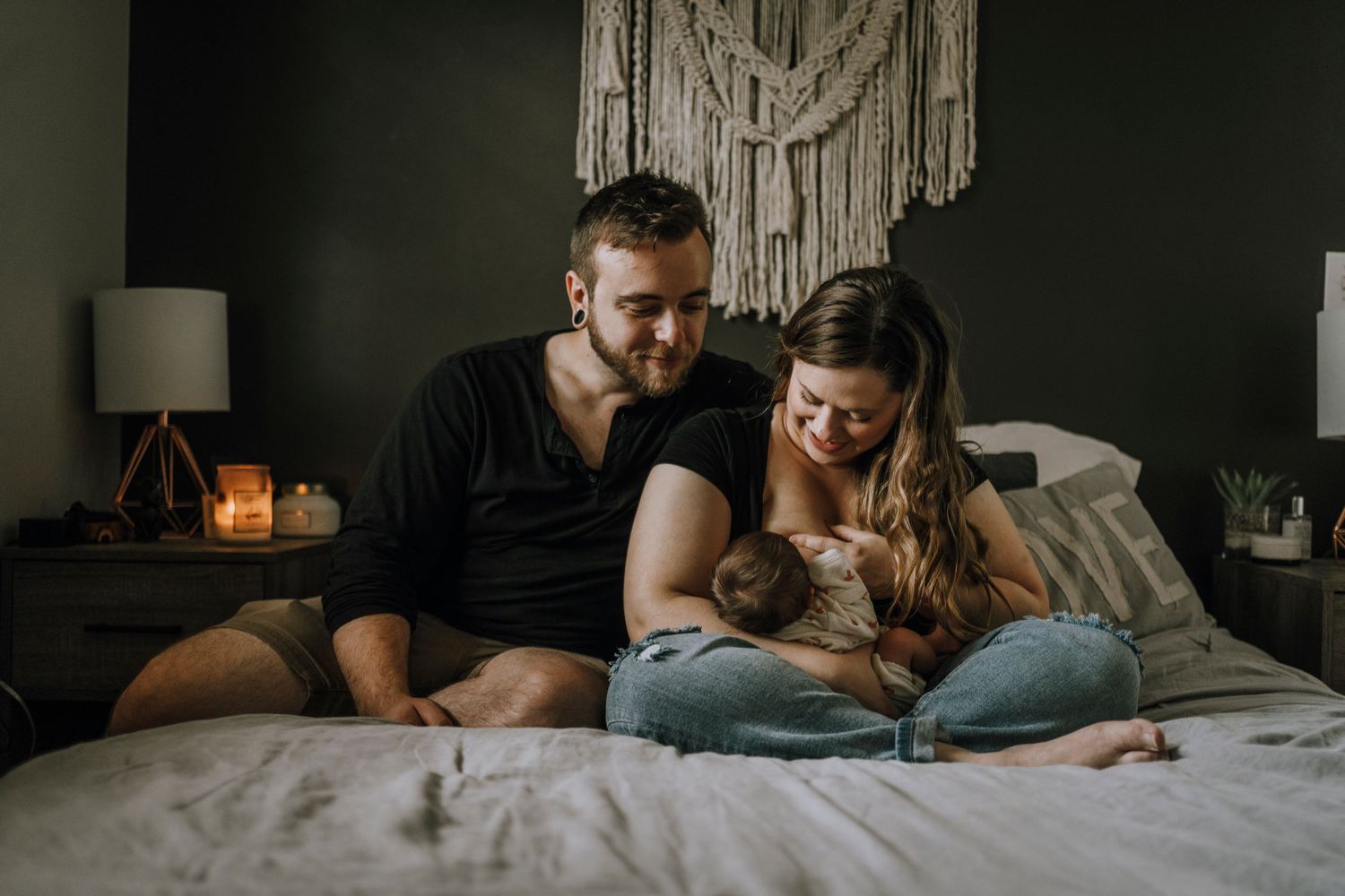 A couple snuggles with their pet dog on a bed with a boho wall decoration.